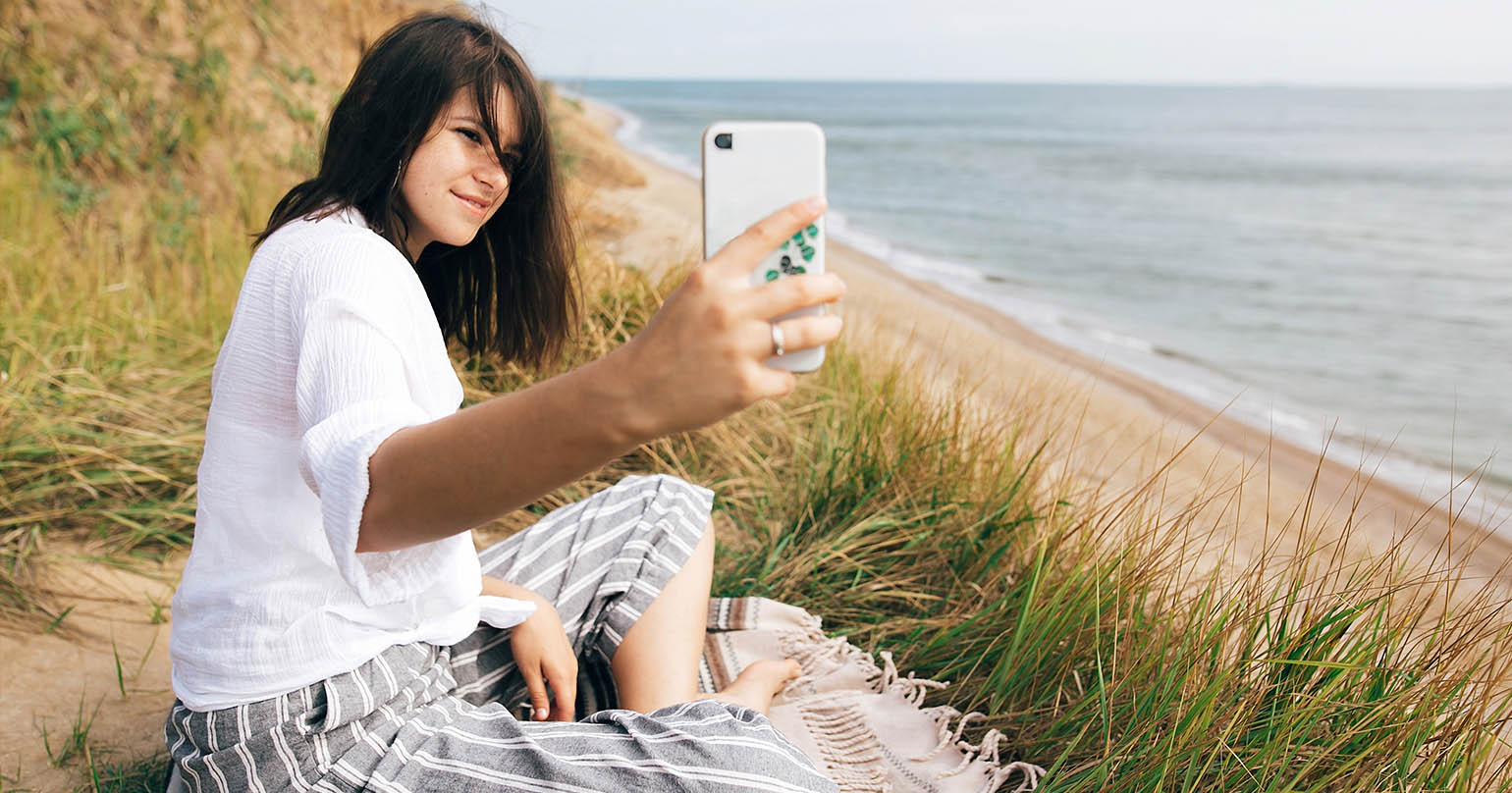 ciemnowłosa kobieta siedząca na kocu na plaży z telefonem w ręku, robiąca sobie zdjęcia do mediów społecznościowych jako symbol samotności w związku